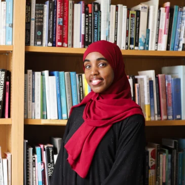 Najmo Ahmed, USM's 2026 student commencement speaker, smiles in front of a wall of bookshelves.