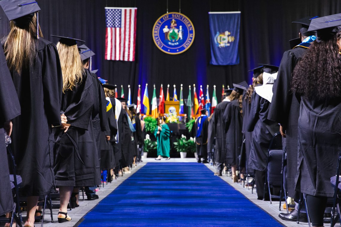 Graduates in black caps and gowns stand on either side of a royal blue aisle leading toward the stage at USM's 146th Commencement ceremony, with the University seal, American flag, and Maine state flag visible in the background.