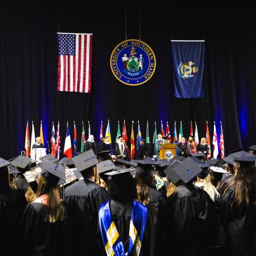 Graduates in black caps and gowns face the stage at Cross Insurance Arena during USM's 146th Commencement ceremony, with the University seal, American flag, and Maine state flag displayed above the stage party.