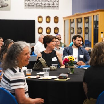 Attendees seated at round tables laugh and chat during the "Good People, Great Connections" networking event at USM's Alumni Reception Center, with championship plaques lining the walls behind them.