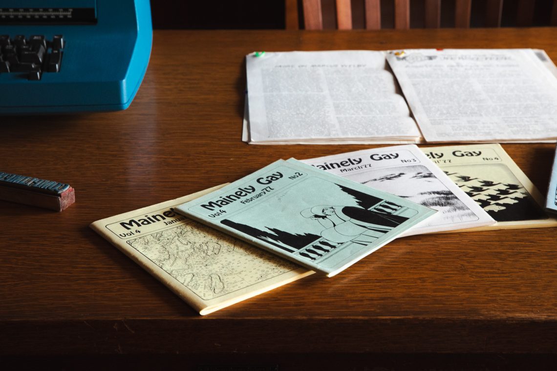 Four early 1977 issues of 'Mainely Gay' newspaper fanned out on a wooden table next to a vintage typewriter and ink stamp, marking the publication's transition from a typed newsletter to a full newspaper.