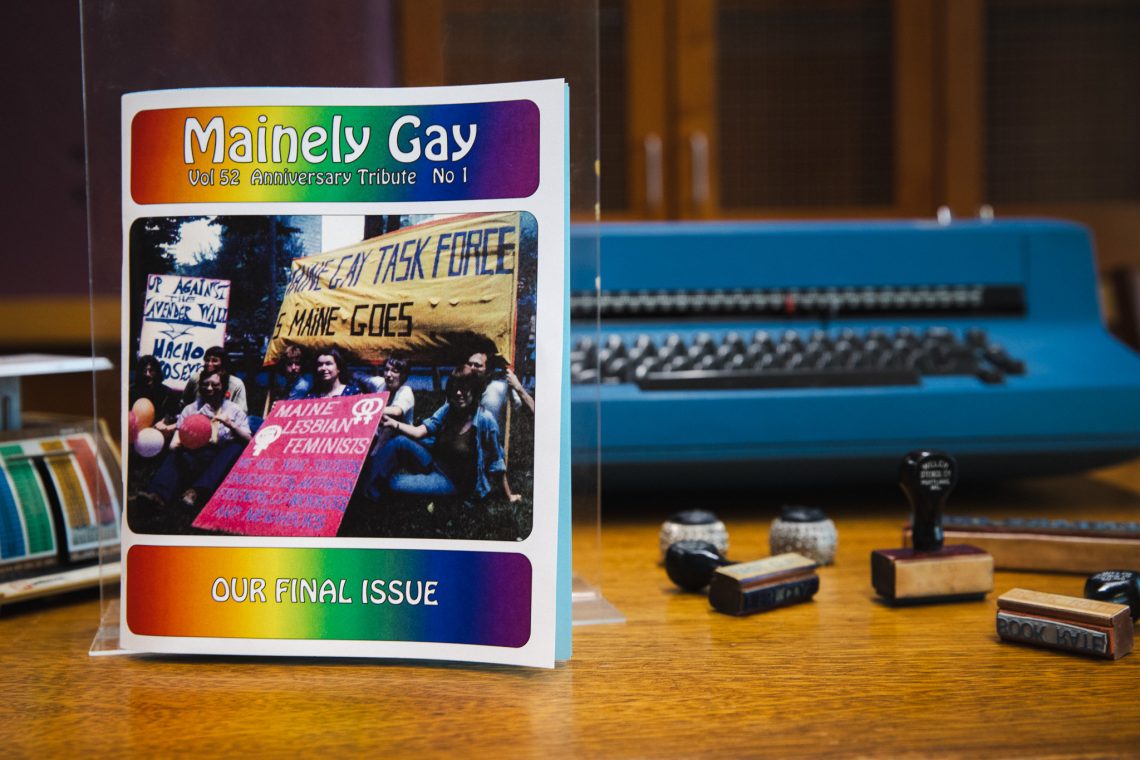 The cover of the 50th anniversary tribute issue of 'Mainely Gay,' displayed next to an original typewriter and stamps used to produce the newsletter in the 1970s.
