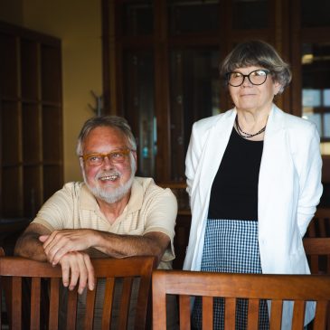 Steve Bull and Susie Bock pose together at USM Special Collections, Bull seated and smiling with his arms resting on a chair, Bock standing beside him.