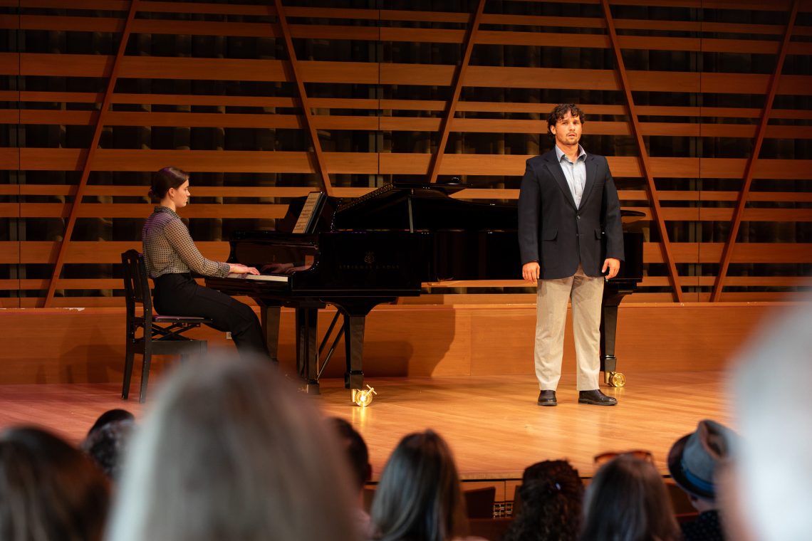 A tenor sings while standing on stage at USM's Crewe Center for the Arts, accompanied by a pianist seated at a grand piano, with an audience seated in the foreground.