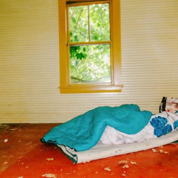 A bare room with a thin mattress on the floor, a blanket and pillow arranged on top, and daylight coming through the room's one window.