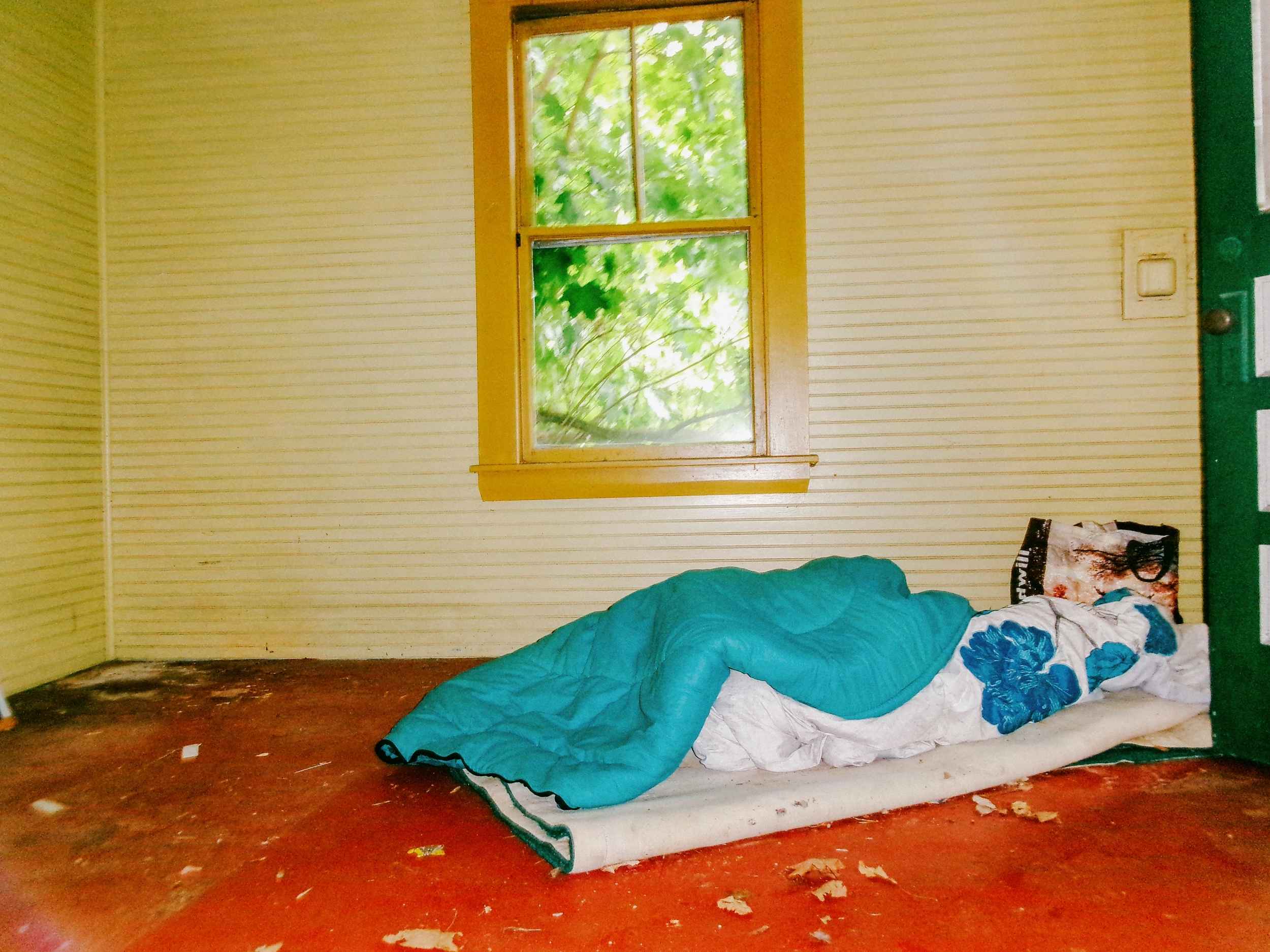 A bare room with a thin mattress on the floor, a blanket and pillow arranged on top, and daylight coming through the room's one window.