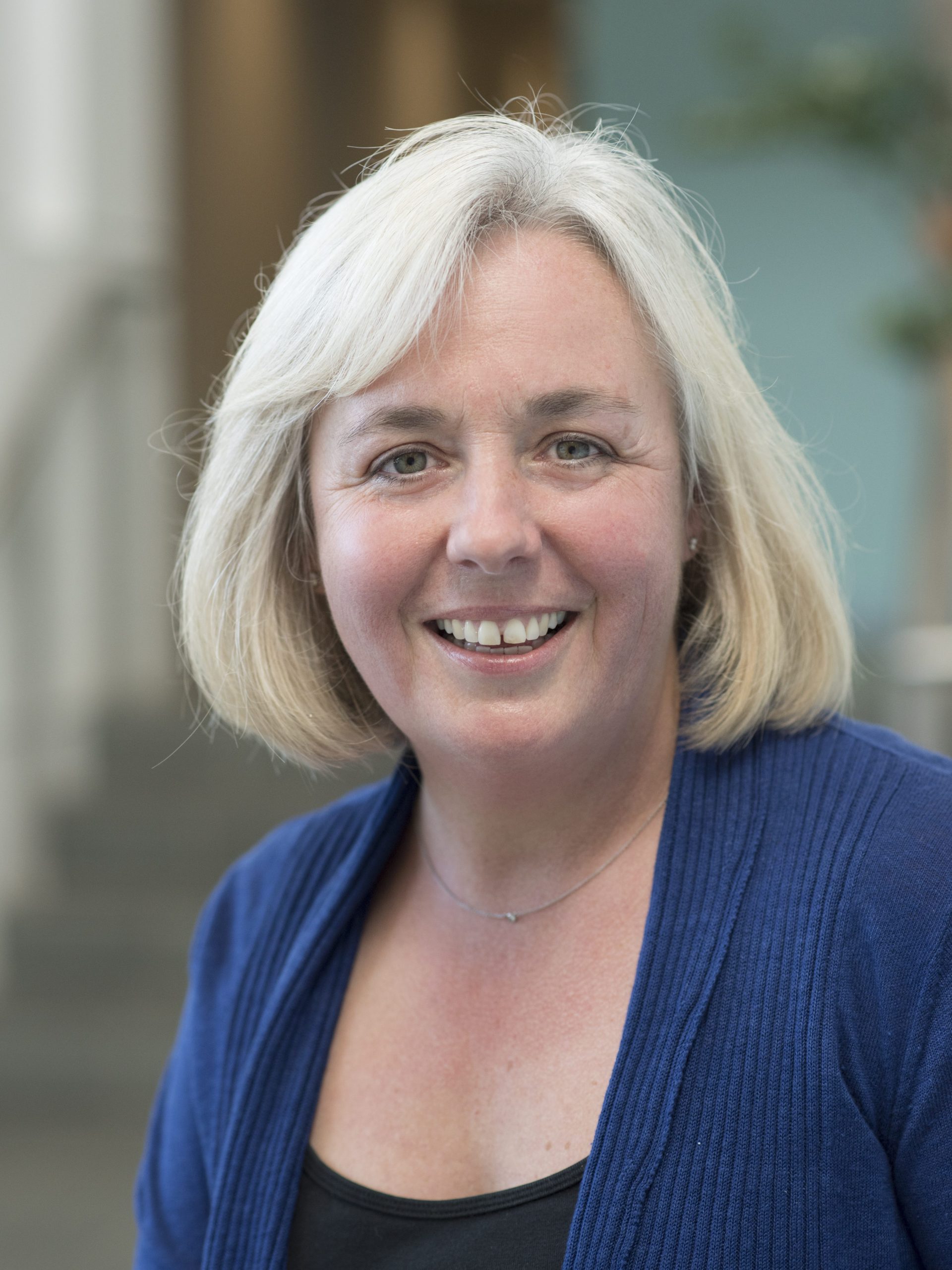 A professional headshot of Cathie Fallona, smiling warmly against a softly blurred indoor background, wearing a navy blue cardigan and a delicate necklace.