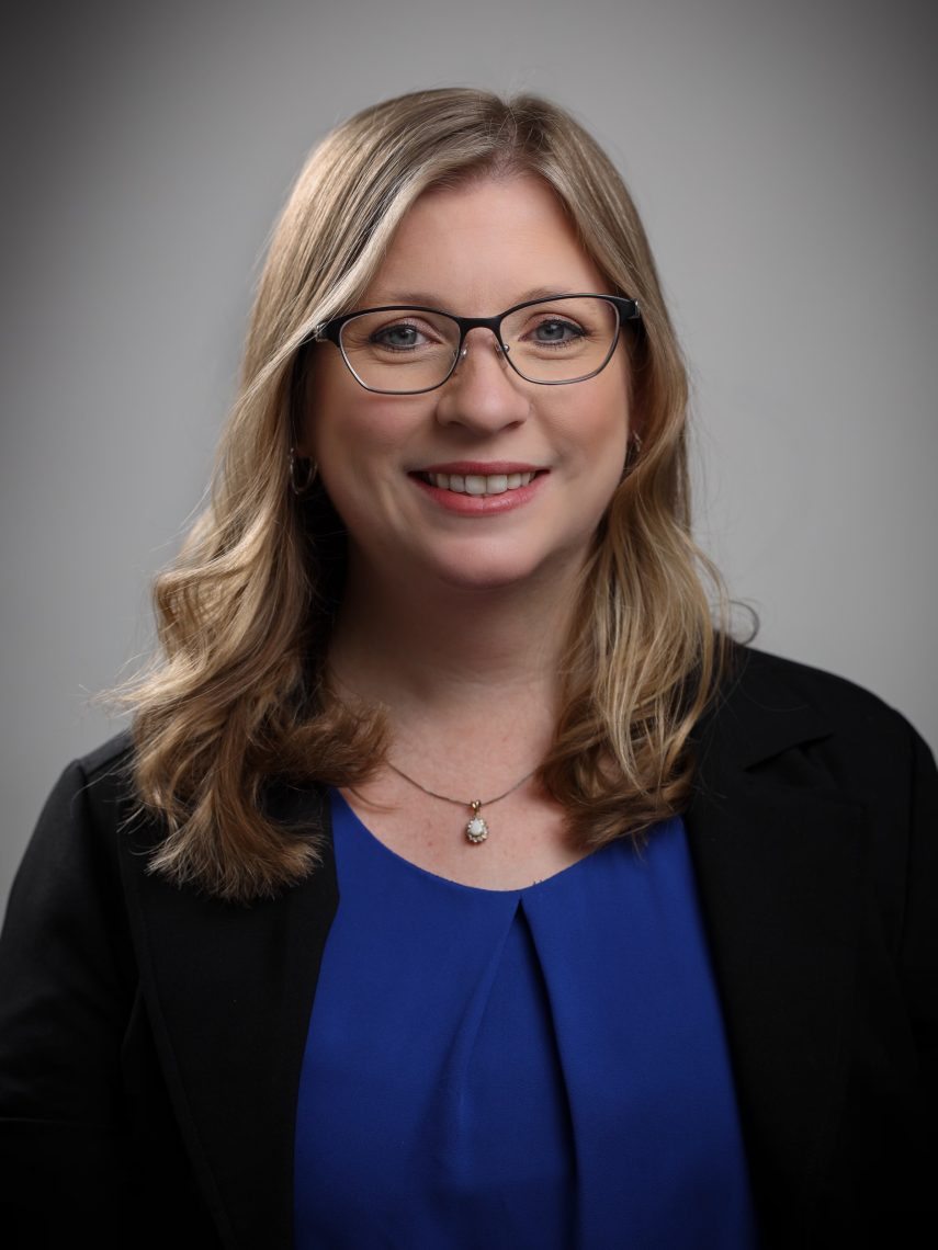 A professional headshot of Kathryn Robinson, smiling against a neutral gray background, wearing a blue blouse, black blazer, and glasses with a delicate pendant necklace.