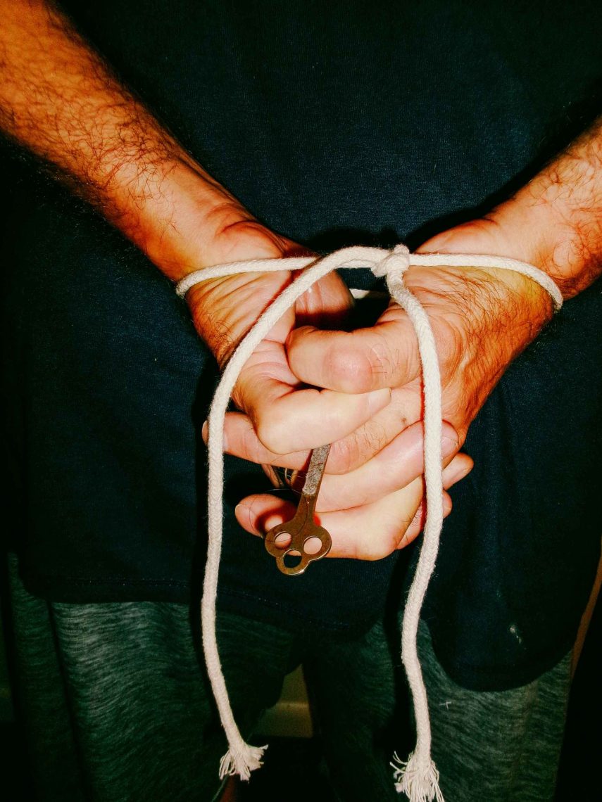 A pair of hands bound behind someone's back with an old fashioned key nestled between their fingers.