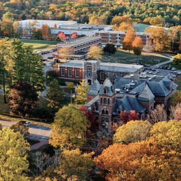 An aerial view of USM's Gorham campus in fall, with brick academic buildings nestled among trees displaying vibrant autumn foliage in shades of orange, red, yellow, and green.