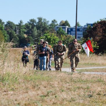 An Army ROTC cadet carries a red and white 'ROTC B' company flag while leading a group of cadets in uniform and civilians in '9/11 Memorial Ruck' shirts along a gravel path through tall grass near Back Cove.