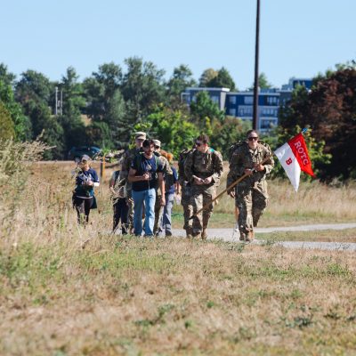 An Army ROTC cadet carries a red and white 'ROTC B' company flag while leading a group of cadets in uniform and civilians in '9/11 Memorial Ruck' shirts along a gravel path through tall grass near Back Cove.