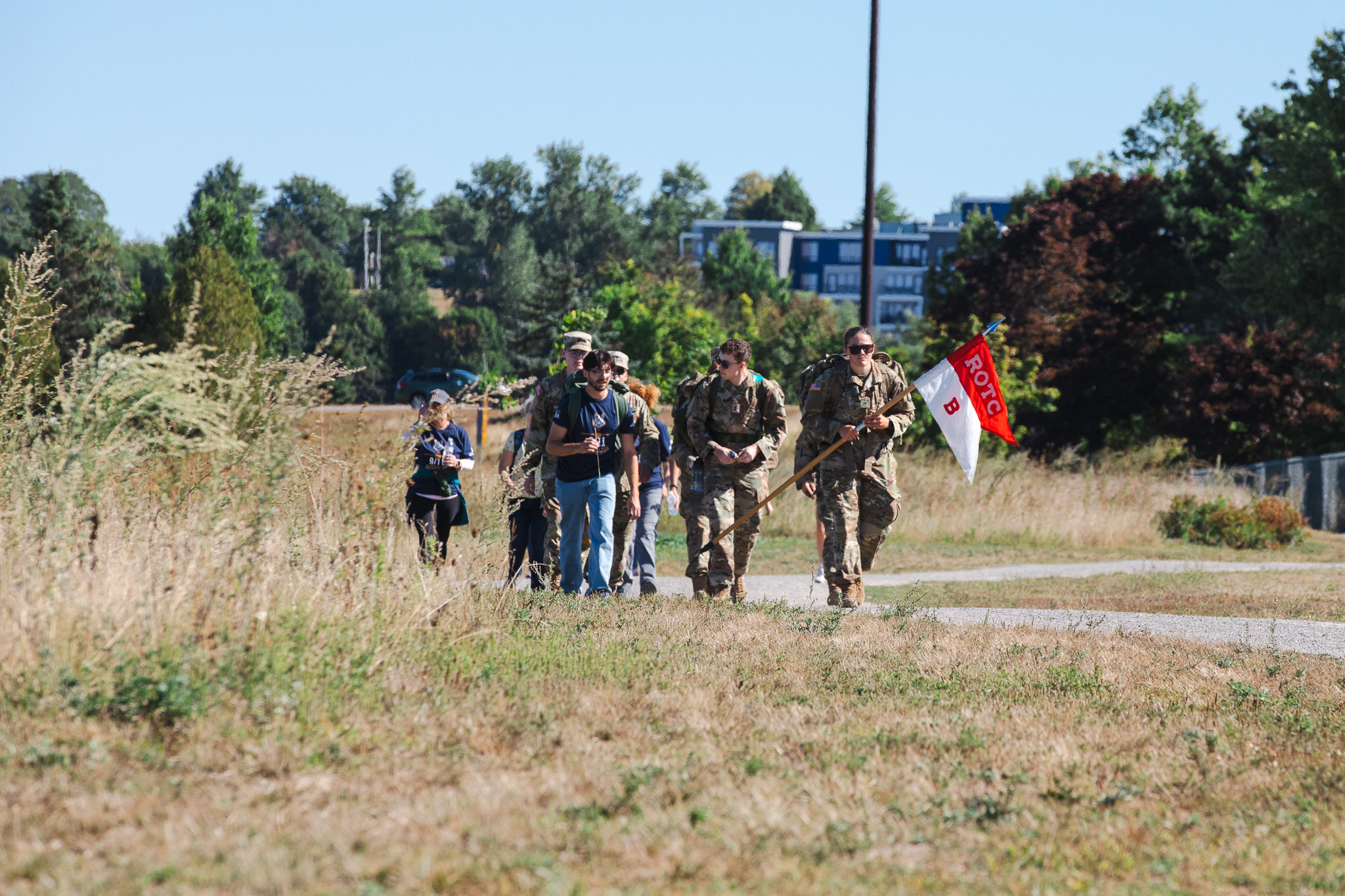 An Army ROTC cadet carries a red and white 'ROTC B' company flag while leading a group of cadets in uniform and civilians in '9/11 Memorial Ruck' shirts along a gravel path through tall grass near Back Cove.