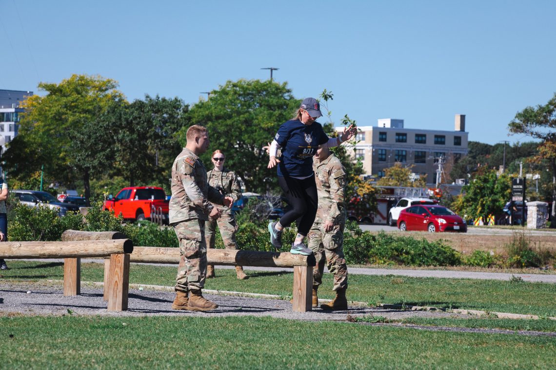 A woman in a navy '9/11 Memorial Ruck' T-shirt jumps down from a low wooden balance beam on an obstacle course, arms out for balance, as two Army ROTC cadets in uniform stand nearby.