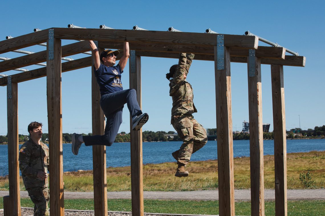A participant in a '9/11 Memorial Ruck' T-shirt swings across monkey bars on an obstacle course beside an Army ROTC cadet in uniform, with Back Cove's water visible behind them and another cadet watching from below.