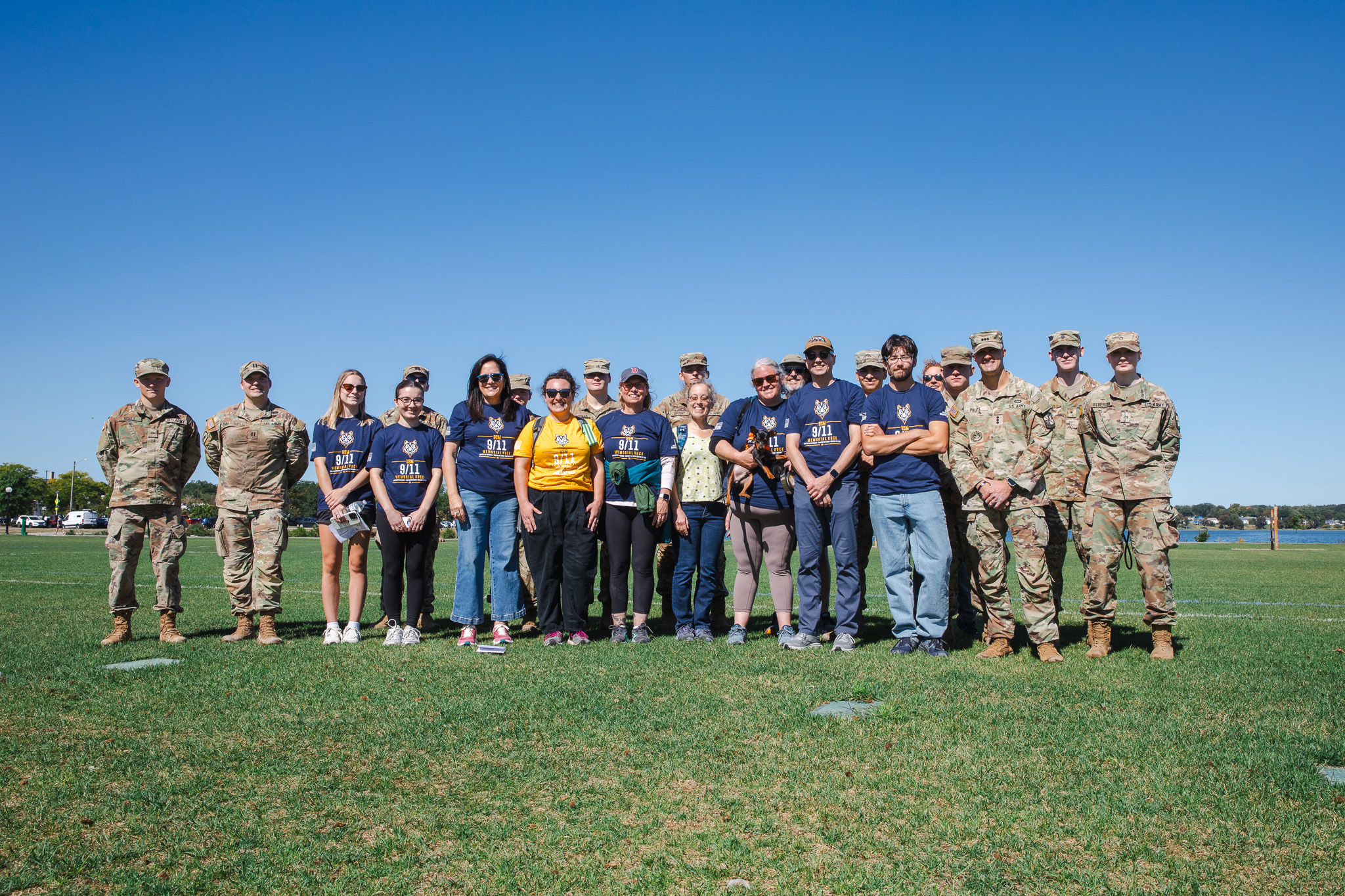 A group of about 20 people, including USM Army ROTC cadets in uniform and civilians wearing navy '9/11 Memorial Ruck' T-shirts, pose together on a grassy field near Back Cove under a clear blue sky.