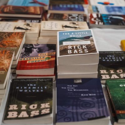 A table covered with stacked copies of books by author Rick Bass, including "The Ninemile Wolves," "Nashville Chrome," "For a Little While," "The Sky The Stars The Wilderness," and "Colter," displayed at a Stonecoast MFA event.