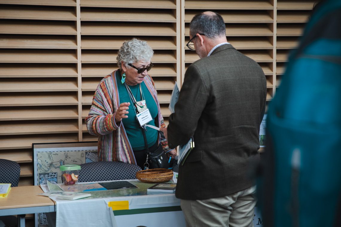 A woman with curly gray hair, sunglasses, and a striped multicolored cardigan gestures as she talks with a man in a brown tweed jacket across a table. The table holds maps, books, and a woven basket, with a framed historical map propped against a chair behind it.