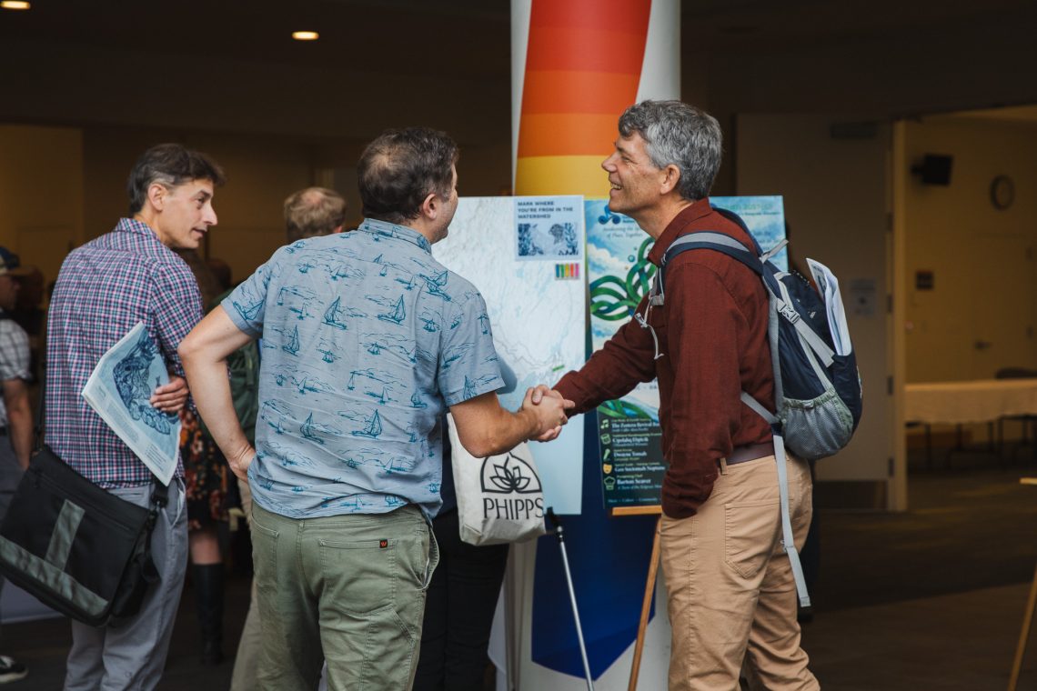 Two men shake hands and smile in a busy event lobby, one wearing a blue shirt printed with sailboats and the other a rust-colored button-up and backpack. Behind them, a large map invites attendees to mark where they're from in the watershed, beside an event poster with a blue and green design. Another attendee nearby holds a printed State of Casco Bay summary.