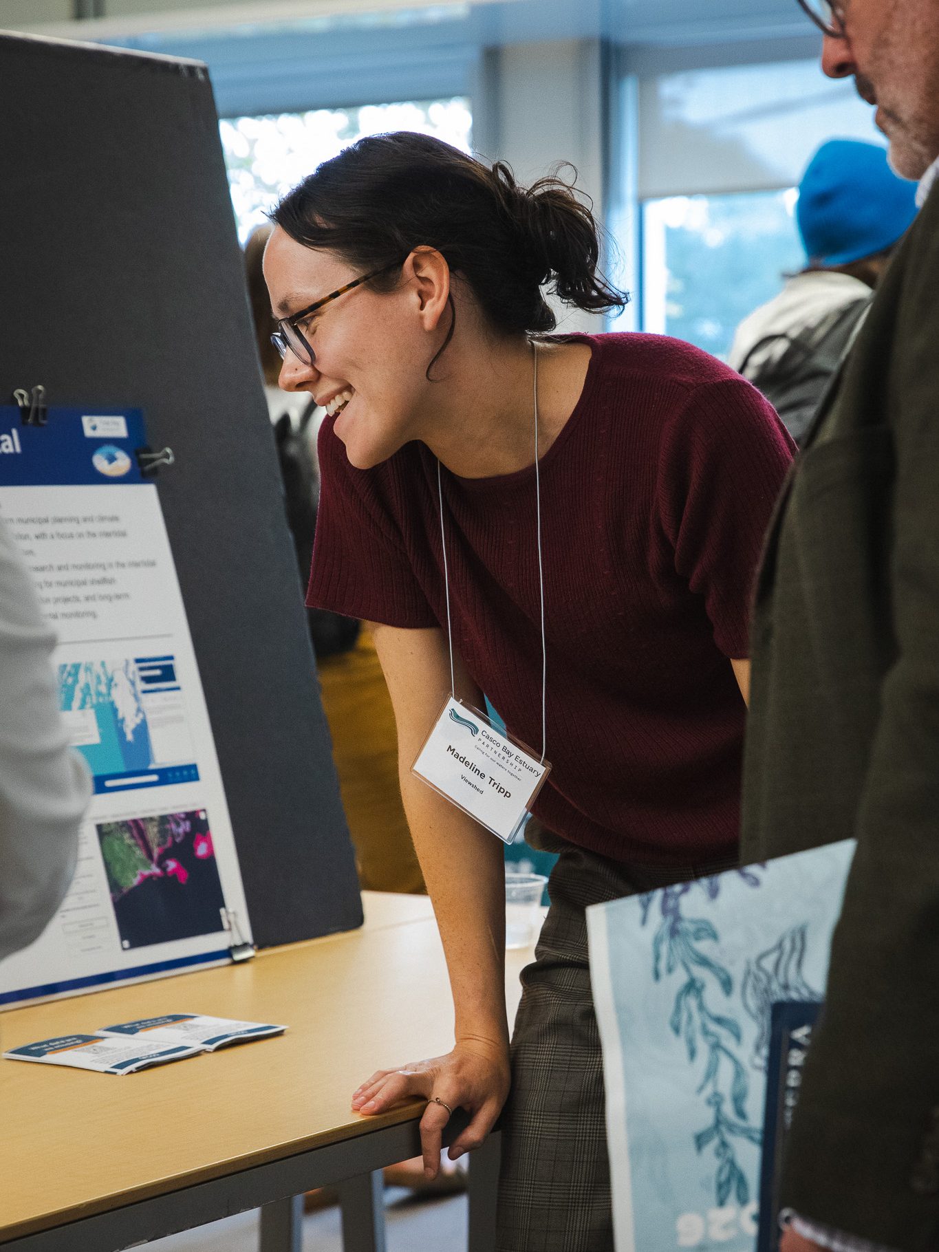 A smiling woman with glasses and a dark ponytail, wearing a maroon knit top, leans against a table as she talks with attendees beside a research poster featuring coastal maps and satellite imagery. A man in the foreground holds a printed State of Casco Bay summary with a blue seaweed illustration.