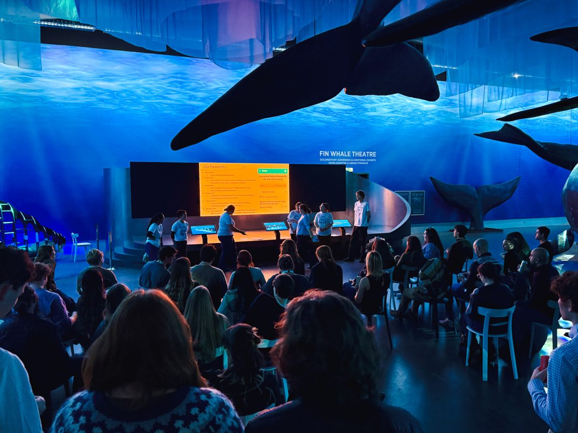 Members of the International Graduate Student Research Cohort present their research on the Arctic blue economy to a seated audience at the Whales of Iceland museum, with life-size whale models suspended overhead.