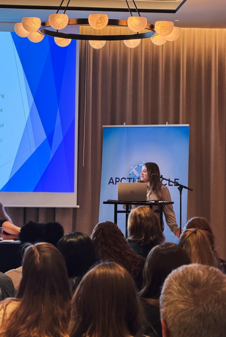 A presenter speaks at a podium in front of an Arctic Circle banner during a session at the Arctic Circle Assembly in Reykjavik, with audience members seated in the foreground.