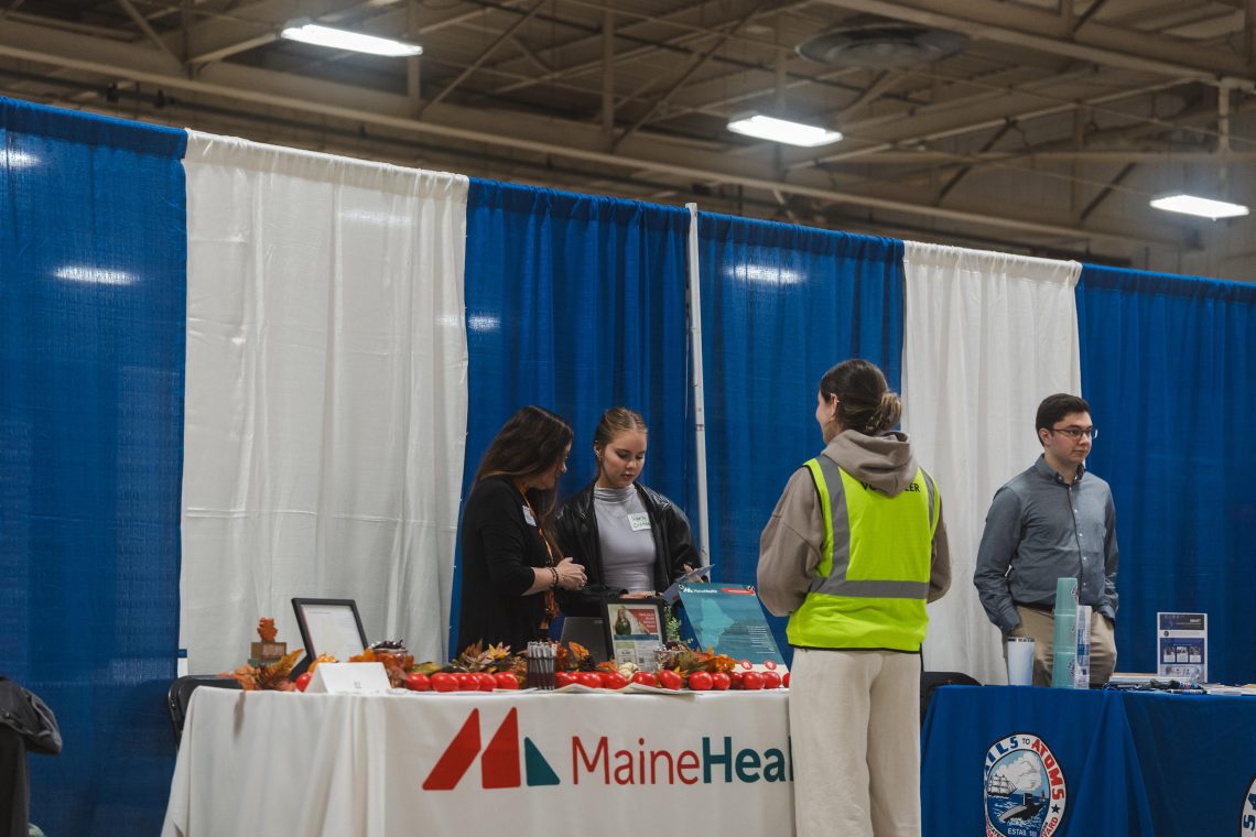 A volunteer in a bright yellow vest speaks with two representatives at the MaineHealth booth, where the table is decorated with autumn leaves and small red pumpkins. A representative at the neighboring Portsmouth Naval Shipyard table stands to the right.