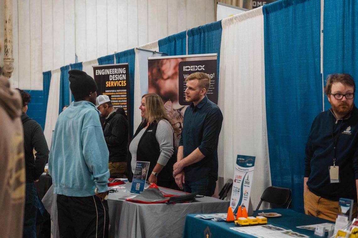 A student in a light blue sweatshirt talks with two IDEXX representatives at their booth during USM’s Job and Internship Fair, as other students and recruiters gather at nearby tables.