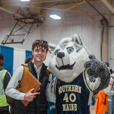 A student in a white button-down shirt and black puffer vest smiles while holding a leather portfolio and posing with Champ, USM’s husky mascot, who wears a navy “Southern Maine” No. 40 jersey. A volunteer in a yellow vest and students carrying bags move between booths in the busy gym behind them.