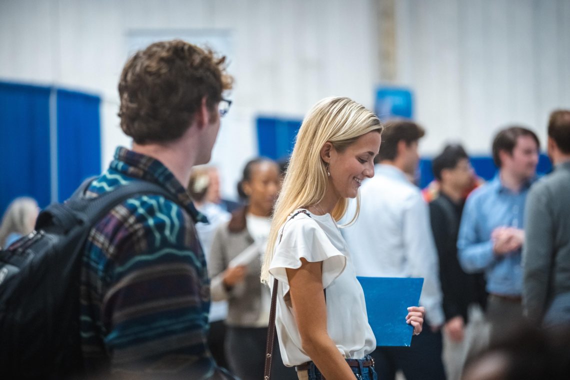 A student in a white blouse smiles as she carries a blue folder past another student with a backpack, while students and recruiters talk at booths in the busy background.