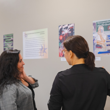 Two visitors talk while viewing photo collage posters by ART 222 students at the Gorham Campus Library.