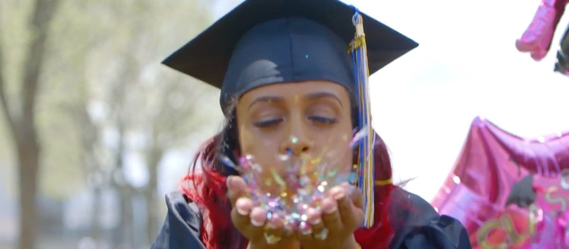 A student wearing a mortarboard with a blue and gold tassel and a graduation gown blows confetti toward the camera. A pink, star-shaped, graduation-themed balloon floats behind her left shoulder.