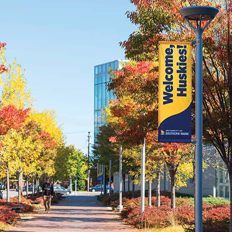 A view of colorful treetops looking down Bedford Street with the Glickman Library in the background. A banner hanging on a lamppost reads 'Welcome Huskies' above the University logo.