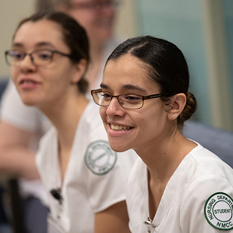 Two students wearing glasses and white scrubs with a Northern Maine Community College (NMCC) patch on the left sleeve.