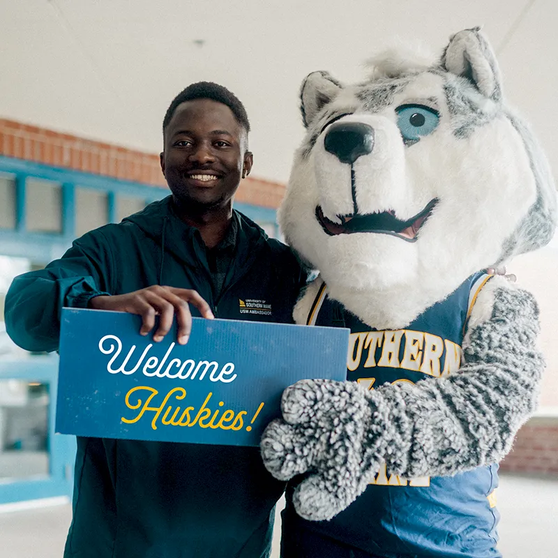 A student and our Husky mascot hold up a welcome sign.