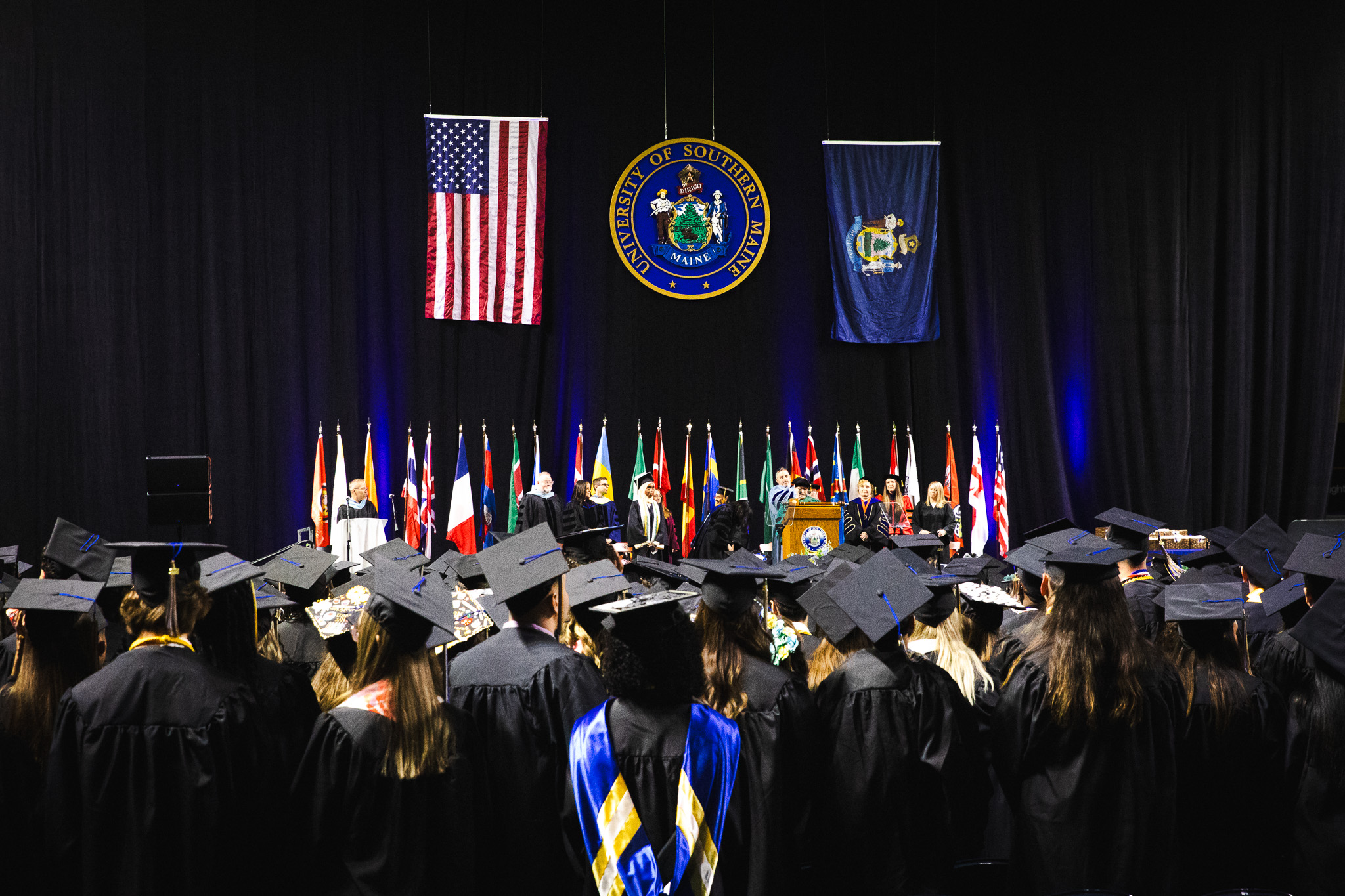 Graduates in black caps and gowns face the stage at Cross Insurance Arena during USM's 146th Commencement ceremony, with the University seal, American flag, and Maine state flag displayed above the stage party.
