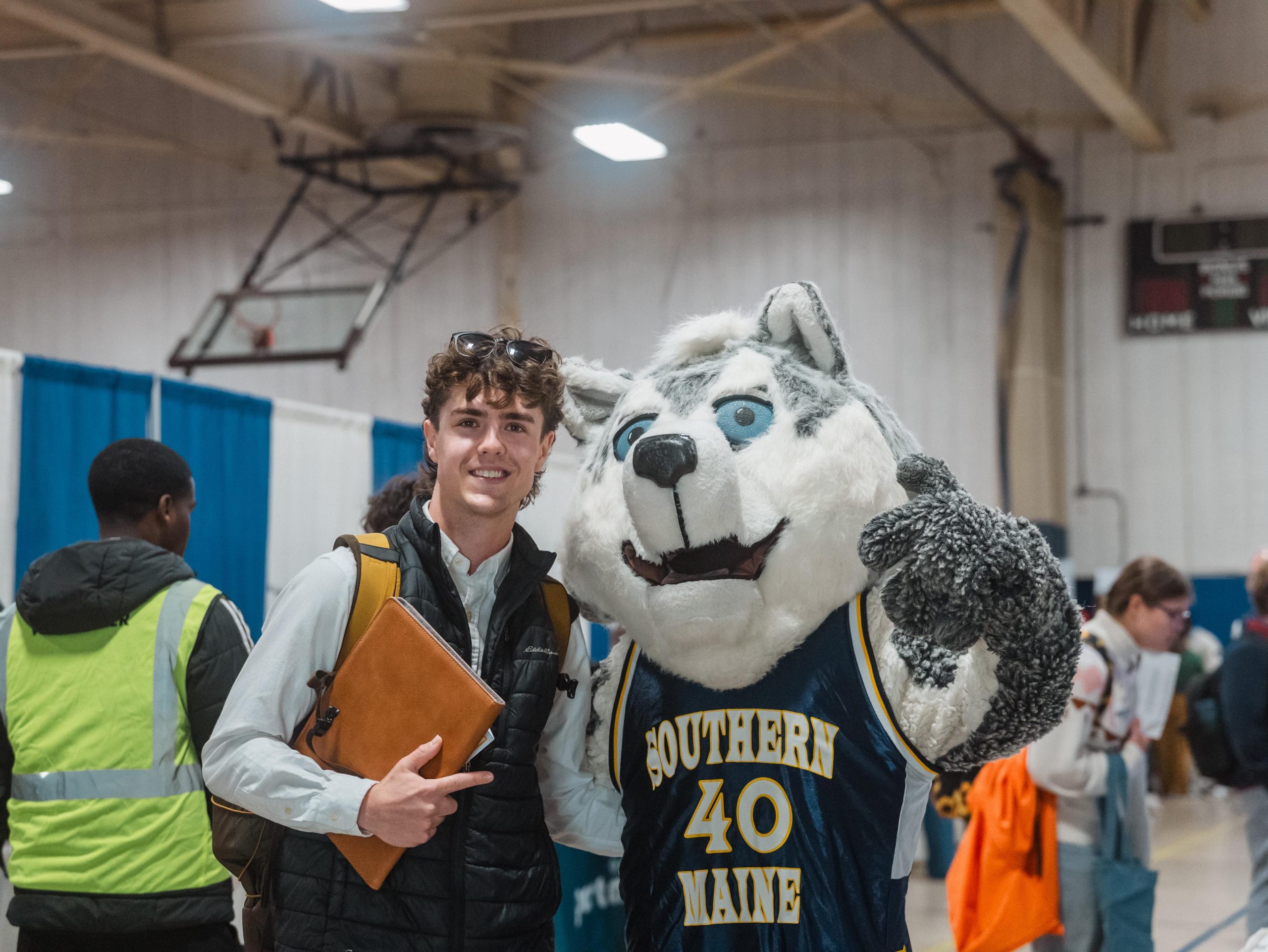 A student in a white button-down shirt and black puffer vest smiles while holding a leather portfolio and posing with Champ, USM’s husky mascot, who wears a navy “Southern Maine” No. 40 jersey. A volunteer in a yellow vest and students carrying bags move between booths in the busy gym behind them.