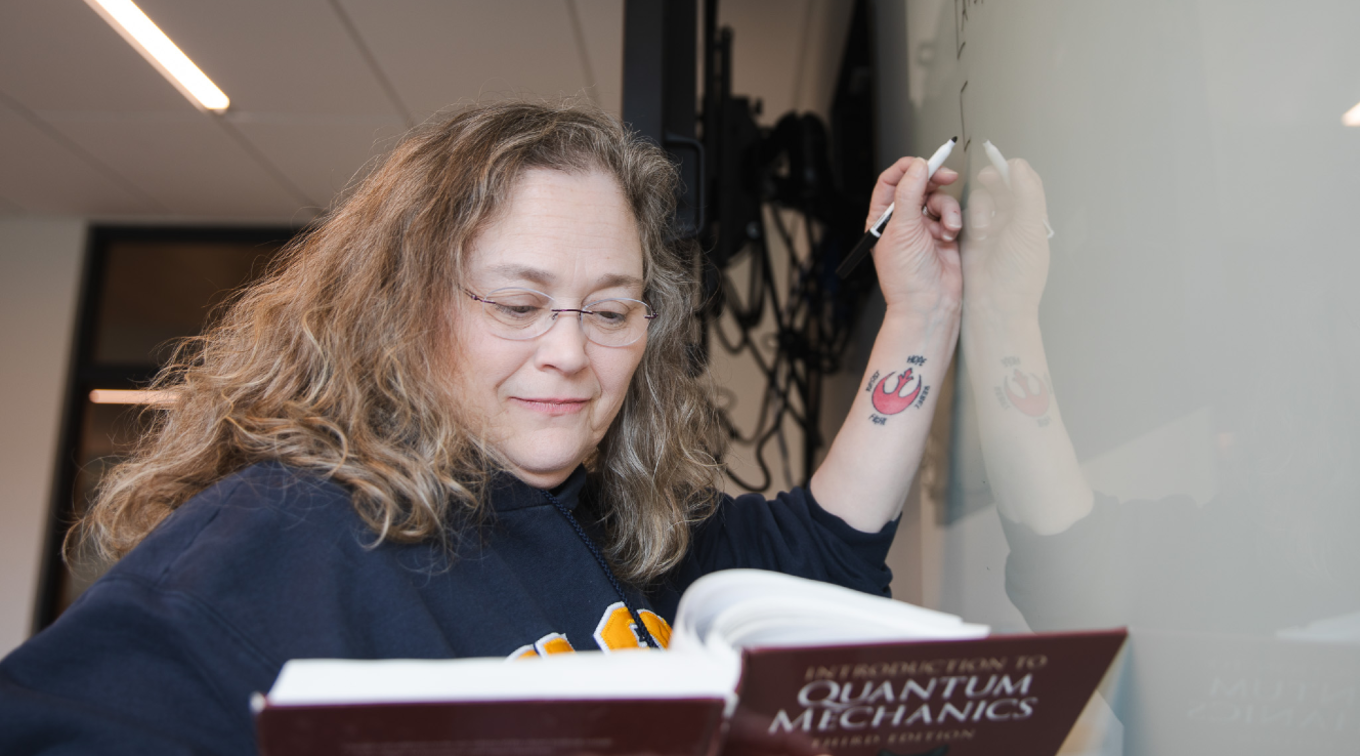 Lisa Struebing references a quantum mechanics textbook while writing an equation on a whiteboard in the Dubyak Center on USM's Portland campus.