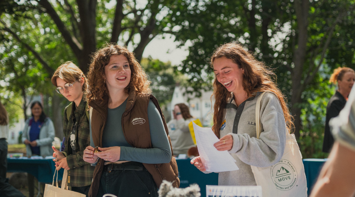 Two students smile and talk at an outdoor table on a sunny day during USM freshman orientation, holding papers in hand.