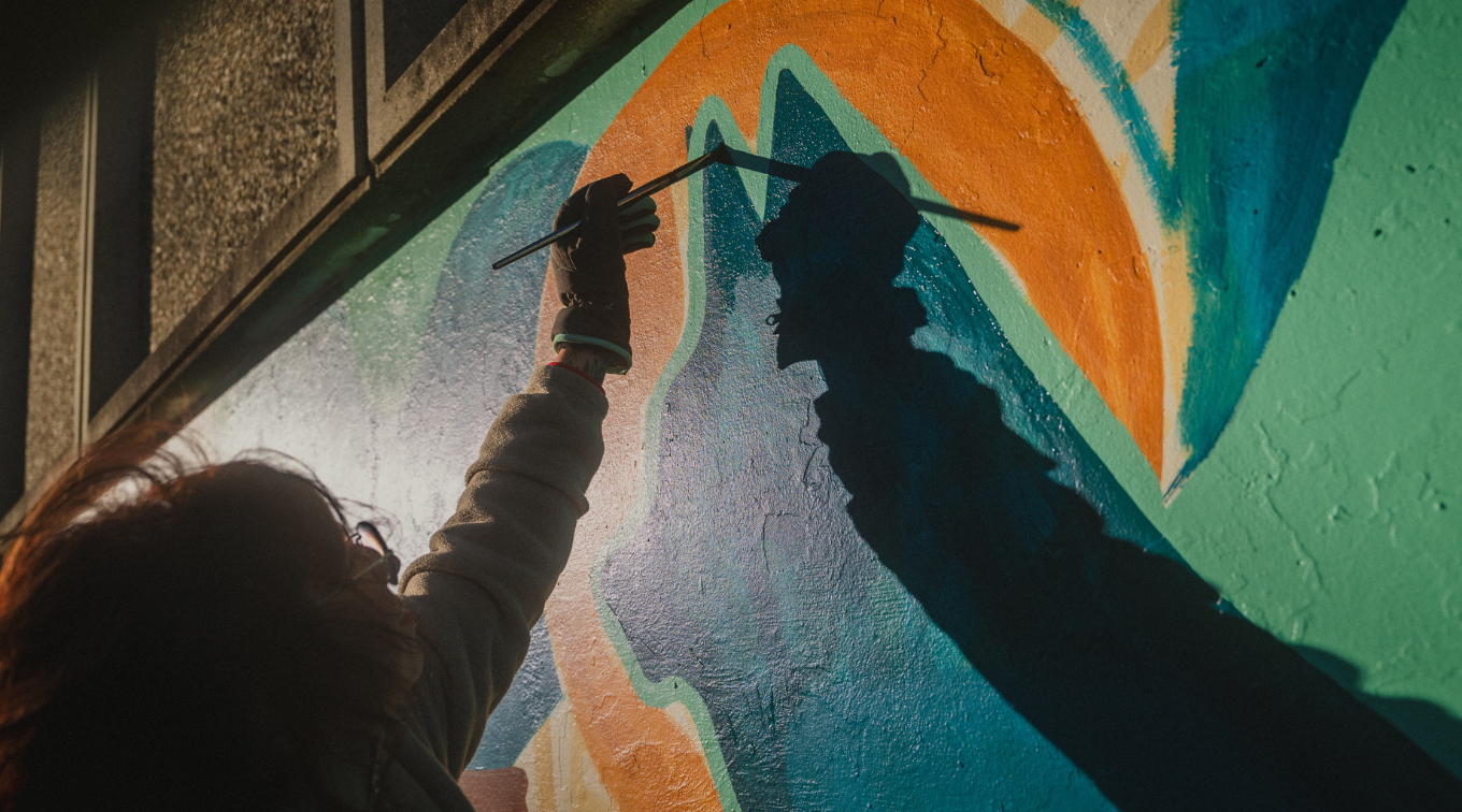 An artist in a work glove reaches up with a paintbrush to add detail to the mural, their shadow stretched across the wall in low sunlight.