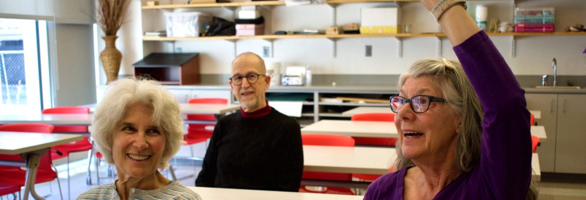 Three OLLI students sit in a classroom. One raises her hand and the others smile.