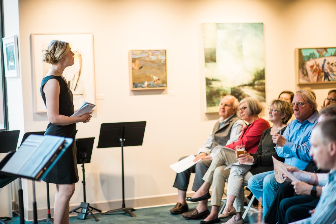 A woman speaks in front of an audience. Musical stands are behind her.