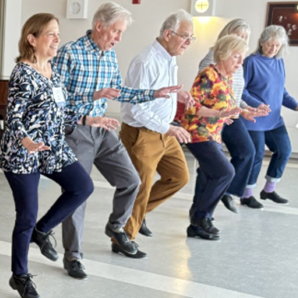 A line of OLLI students tap dance with their left legs up in the air.
