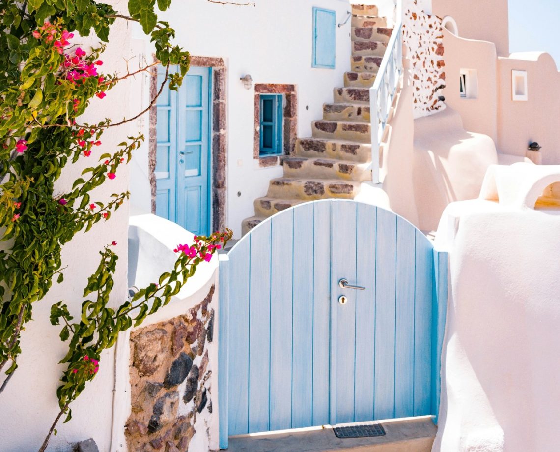 A whitewashed stone house with an open staircase behind a light blue gate in Santorini.