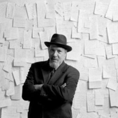 August Wilson in a suit jacket and hat, stands in front of a wall plastered with papers.