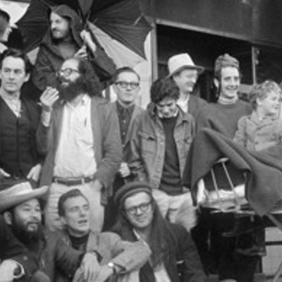 A monochrome photo of a group of diverse people, some holding umbrellas, gather in front of a building.