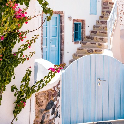 A classic white Greece house with outside stone steps behind a blue garden gate.