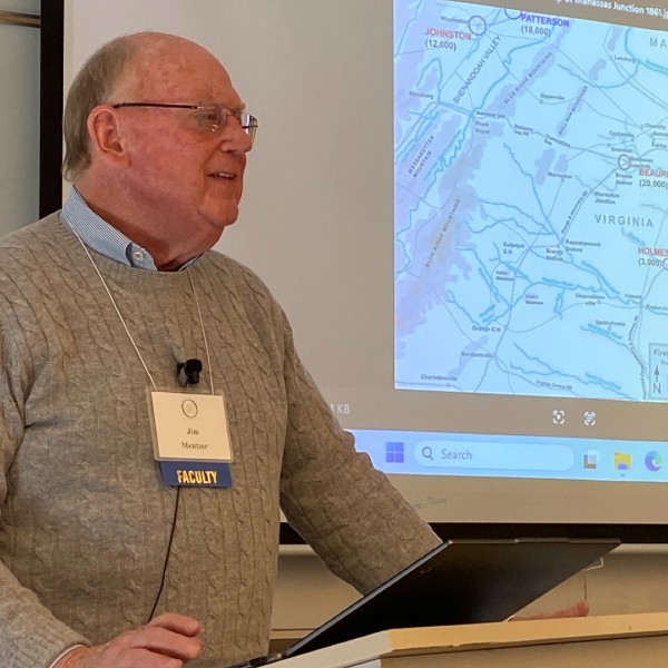 An OLLI instructor stands at a podium with a microphone pinned to his sweater. A map is projected onto a screen behind him.