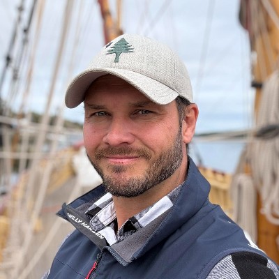 Matt Blazek with a Maine baseball cap on stands in front of sailboat rigging