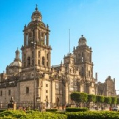 The Metropolitan Cathedral of Mexico City under a clear blue sky, with lush green trees and manicured bushes in the foreground.
